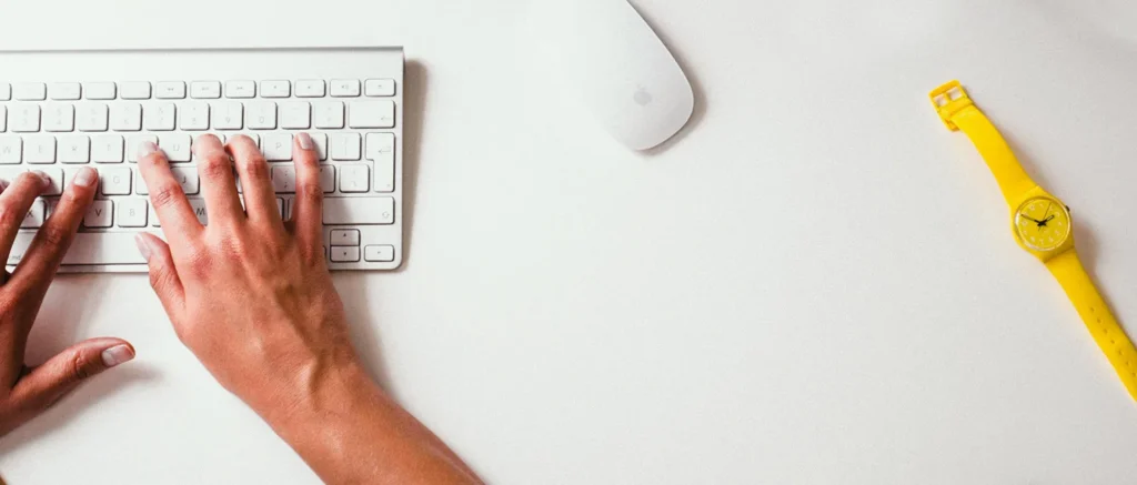 Hands typing on a white keyboard beside a yellow watch