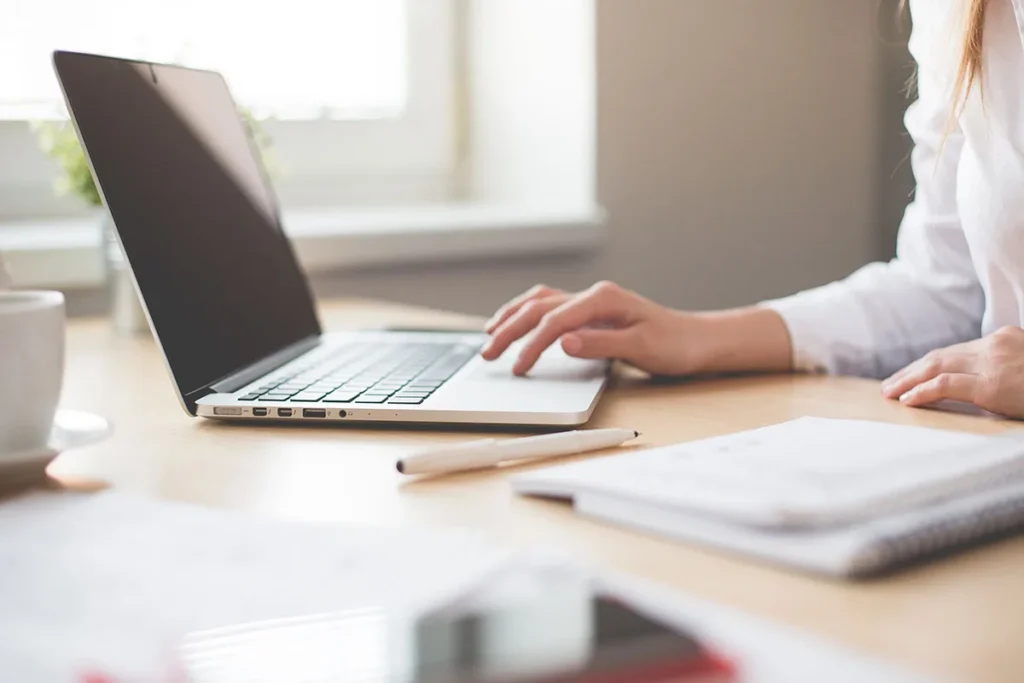 Hands typing on a laptop next to an open notebook