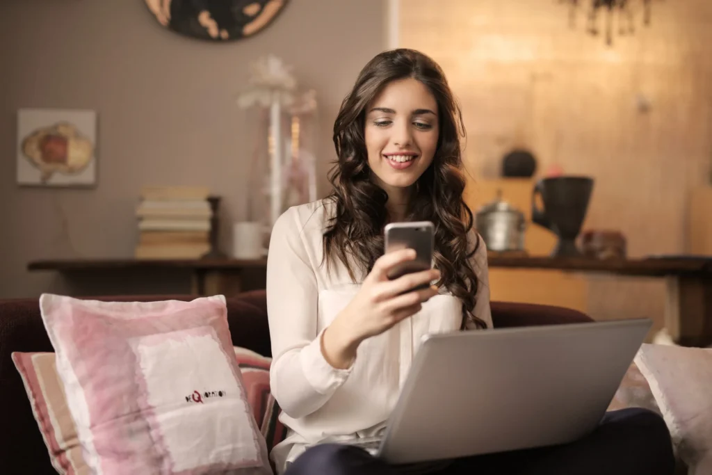 A creator smiling at a phone, with a laptop open on the sofa beside them